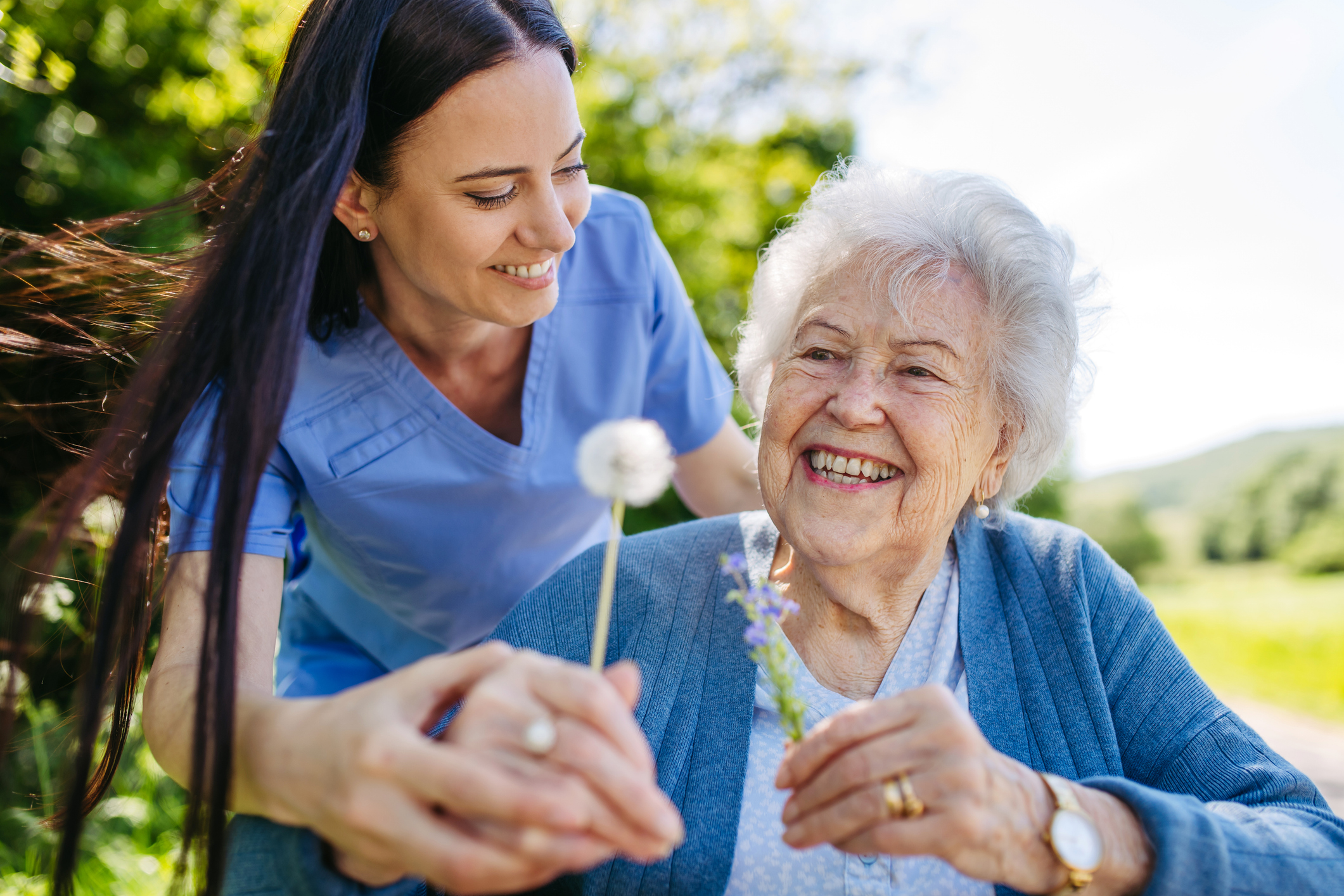 Female caregiver and senior woman in wheelchair holding dandelion, picking wild flowers. Nurse and elderly woman enjoying a warm day in nursing home, public park. Caregiver and senior woman in wheelchair holding dandelion, picking wild flowers. Nurse and elderly woman enjoying a warm day in nursing home, public park.